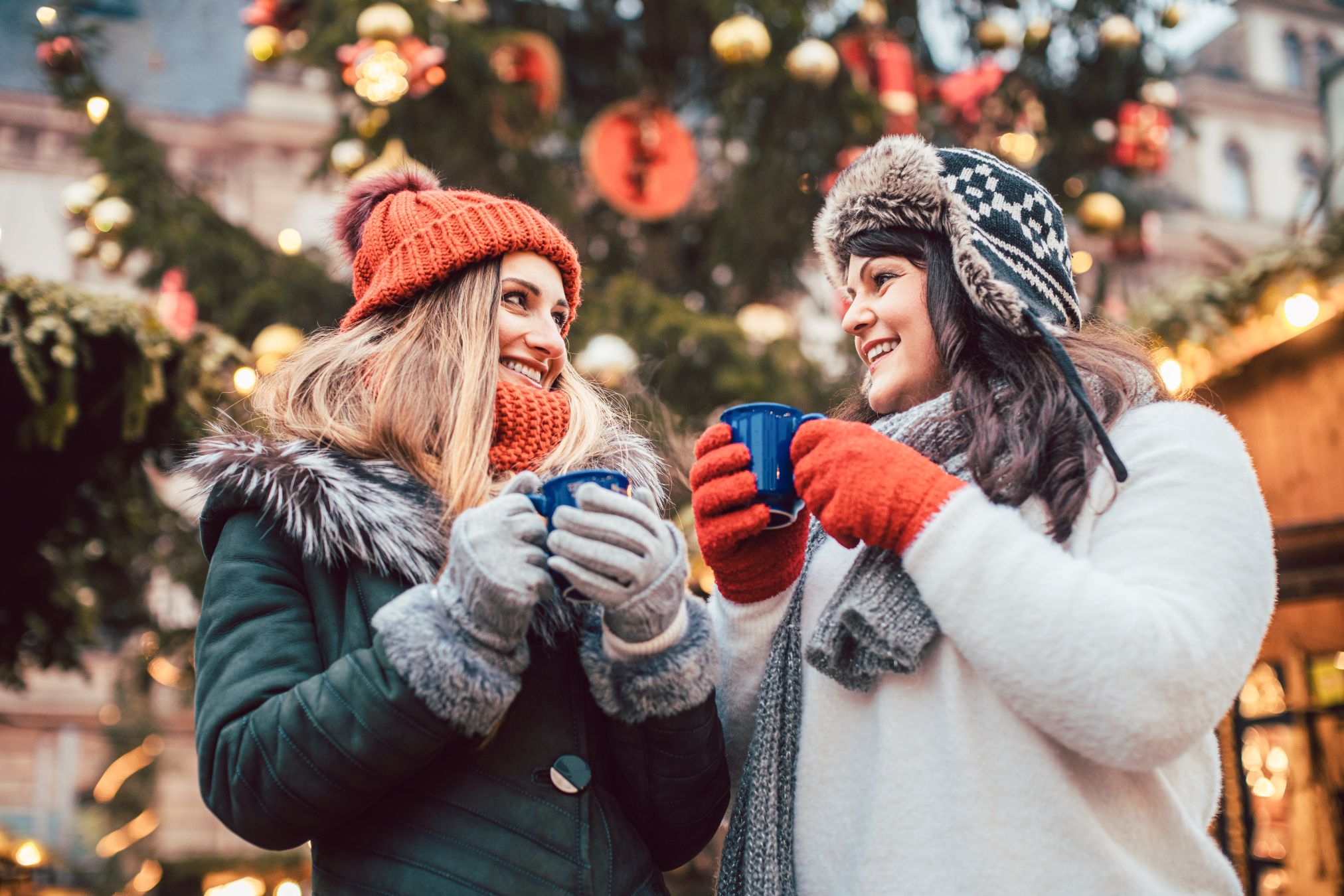 Two women warmly dressed holding mugs in front of Christmas tree